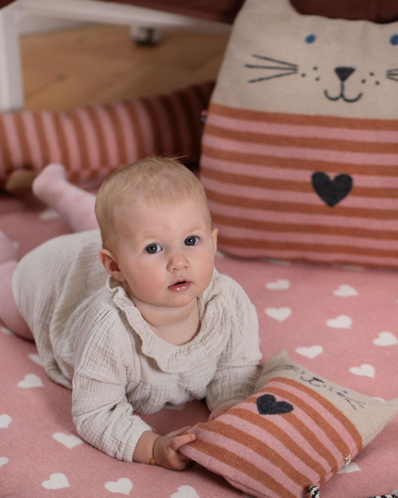 Baby lying on a pink heart-patterned blanket with a striped pillow featuring a cat face.