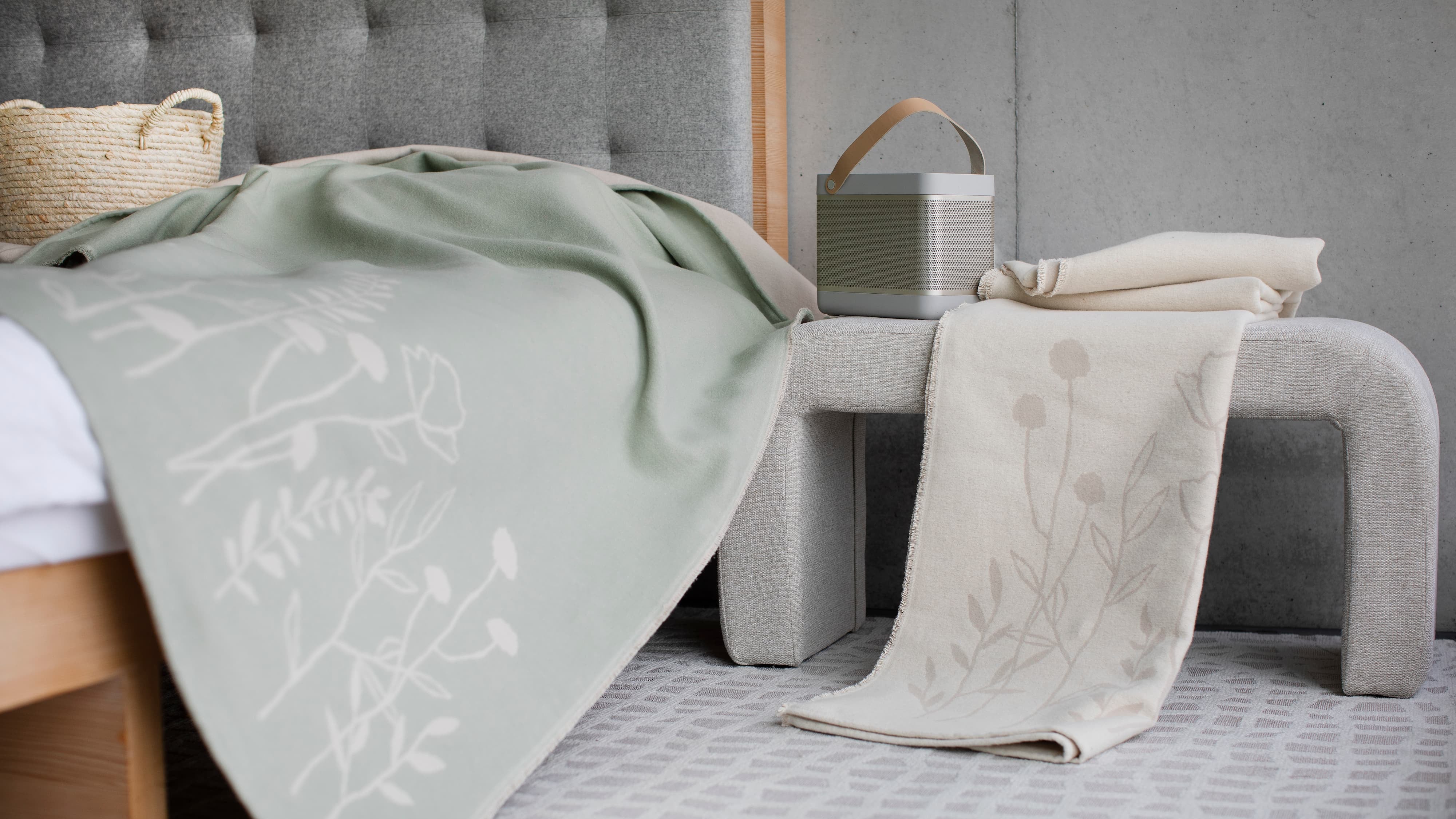 Bed with green blanket and white towel featuring floral patterns, next to a small table with a basket.